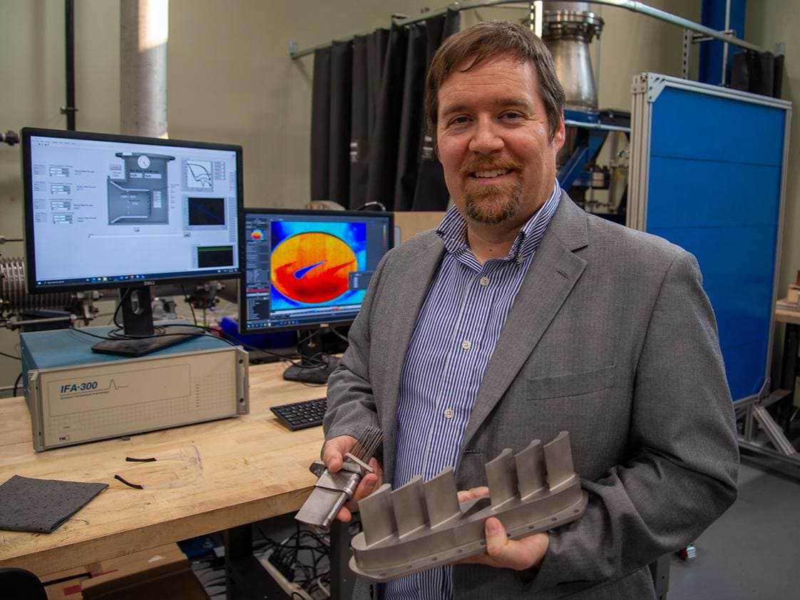Stephen Lynch, associate professor of mechanical engineering, holds metal 3D-printed turbine vanes that are tested in the high speed cascade shown in the background. Lynch was part of a team that 3D printed a turbine component with ceramics, which are more heat tolerant than traditional metals. Credit: Kate Myers/Penn State. All Rights Reserved.